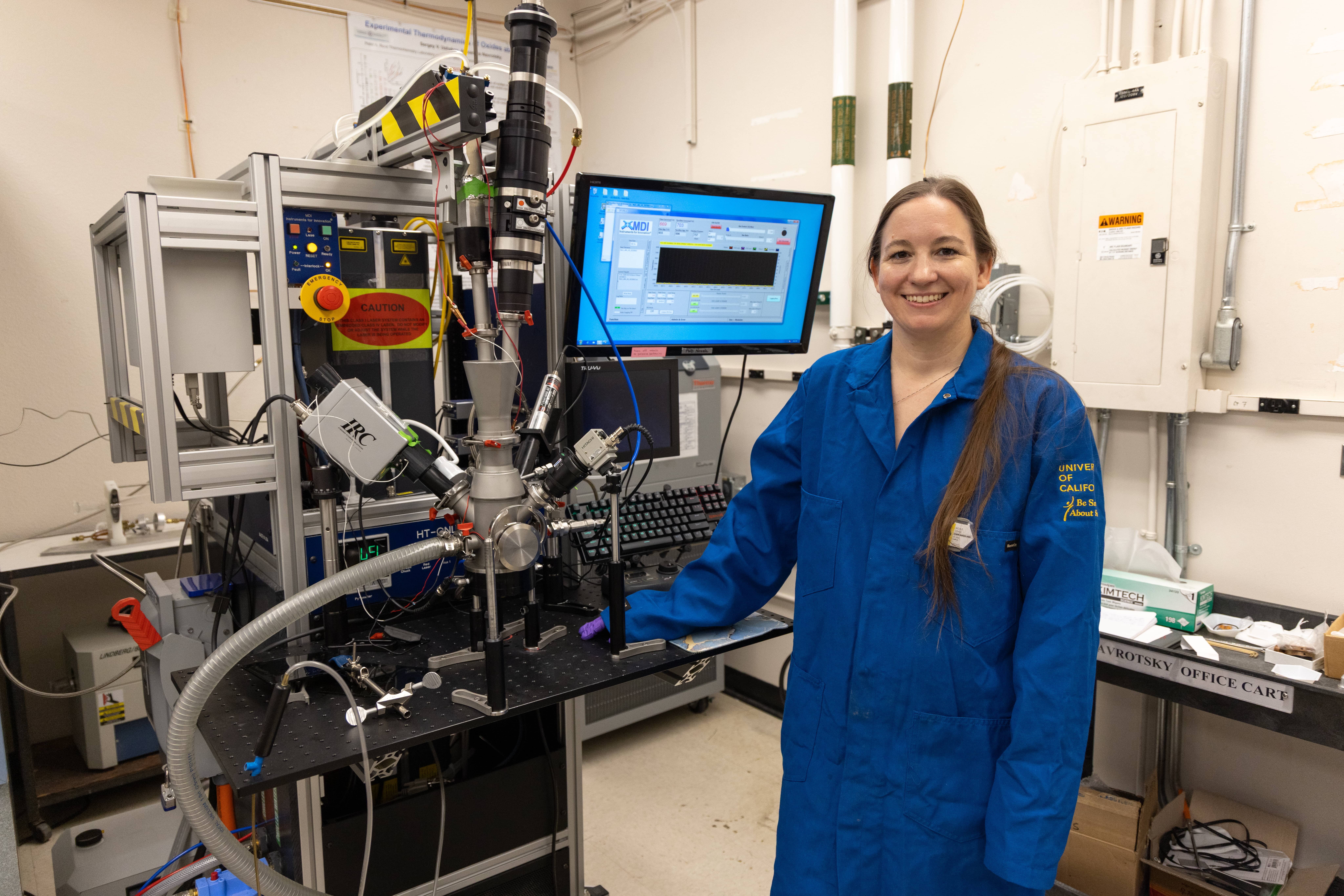 Nicole O'Shea in a blue jumpsuit posing with lab machinery
