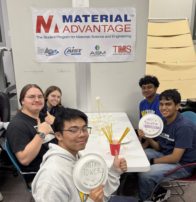 Five students at table with tall spaghetti-and-marshmallow tower under Material Advantage banner