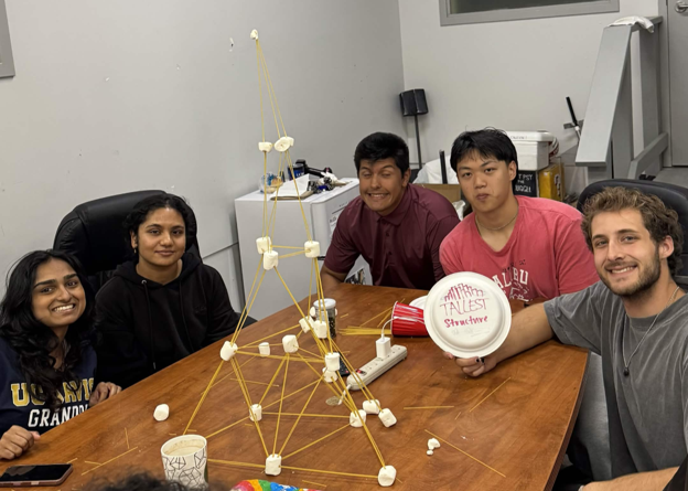 Five students smiling at a table with a tall marshmallow-and-spaghetti tower