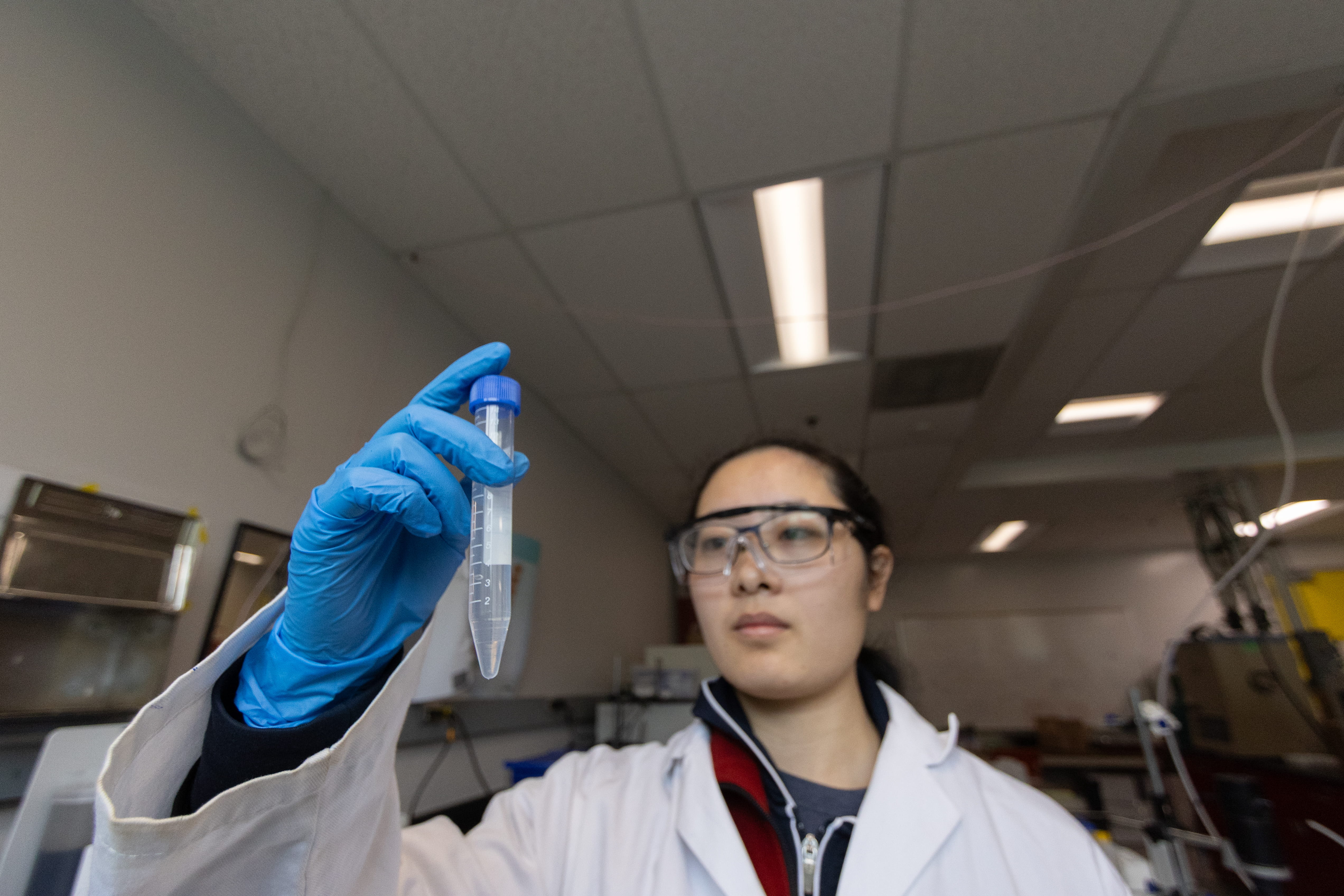 MacAuley in a lab coat and safety glasses examines a test tube in a laboratory.