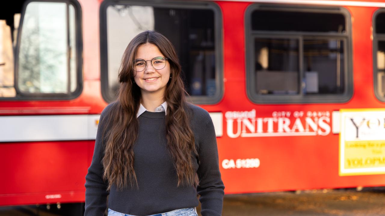 Arden Stratton wears a collared shirt under a sweater and stands outdoors near a red Unitrans bux
