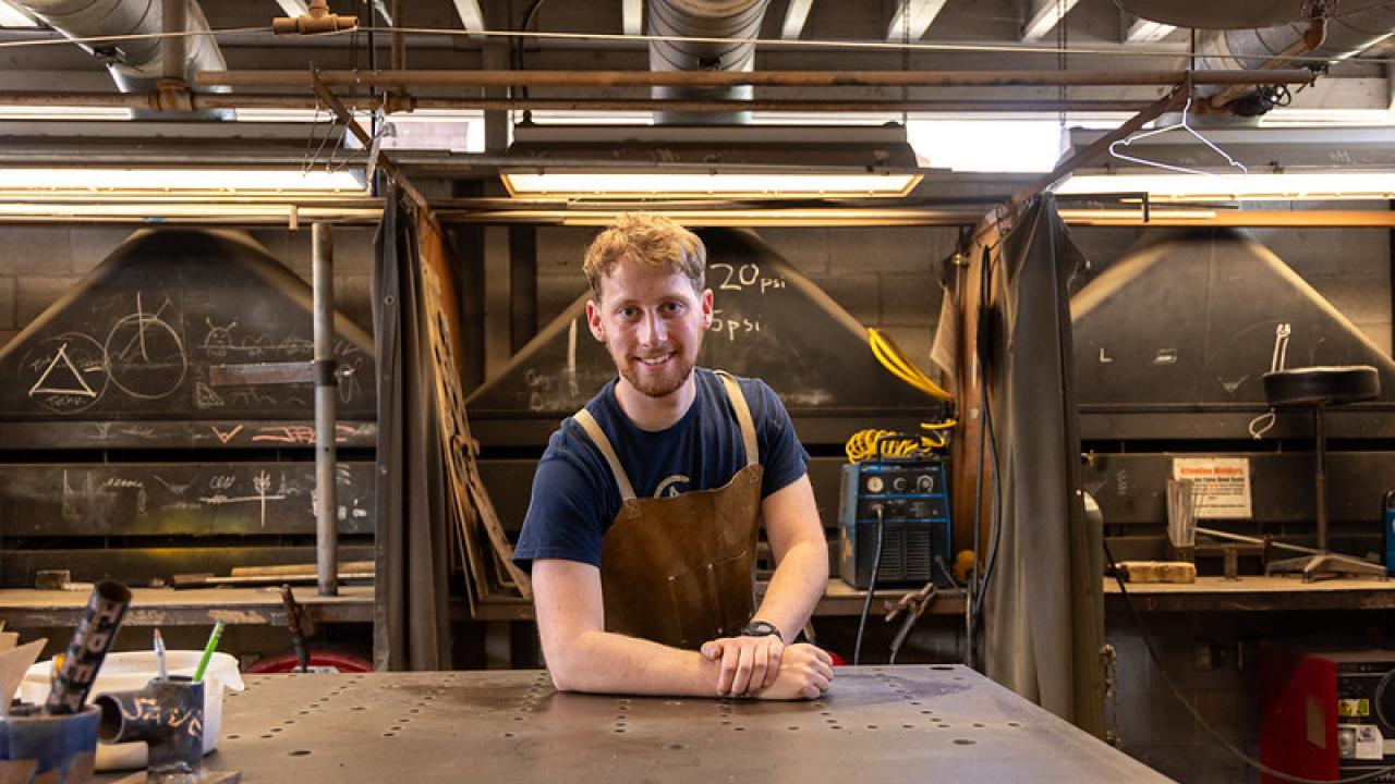 A young man with red hair wears an apron and stands in a welding studio