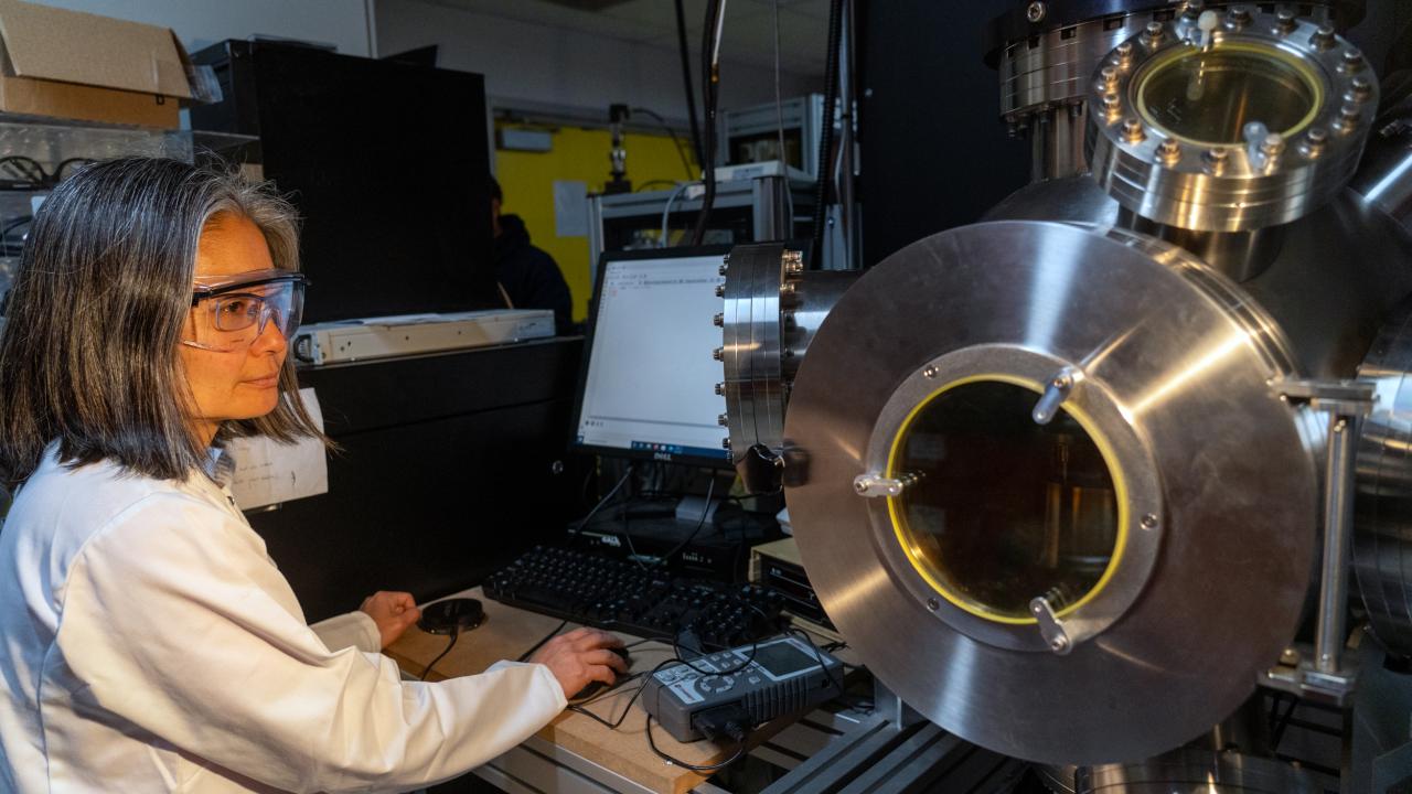 Yayoi Takamura, professor and chair of materials science and engineering wears safety goggles and a lab coat and stands near a machine in her lab