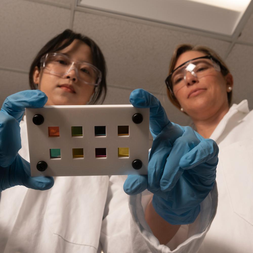 Two researchers stand in a lab looking at a photocell, wearing PPE