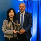 alt="Peifen Lyu stands to the left of Dean Richard Corsi, holding an award in front of a blue backdrop"