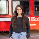 Arden Stratton wears a collared shirt under a sweater and stands outdoors near a red Unitrans bux