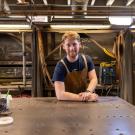 A young man with red hair wears an apron and stands in a welding studio